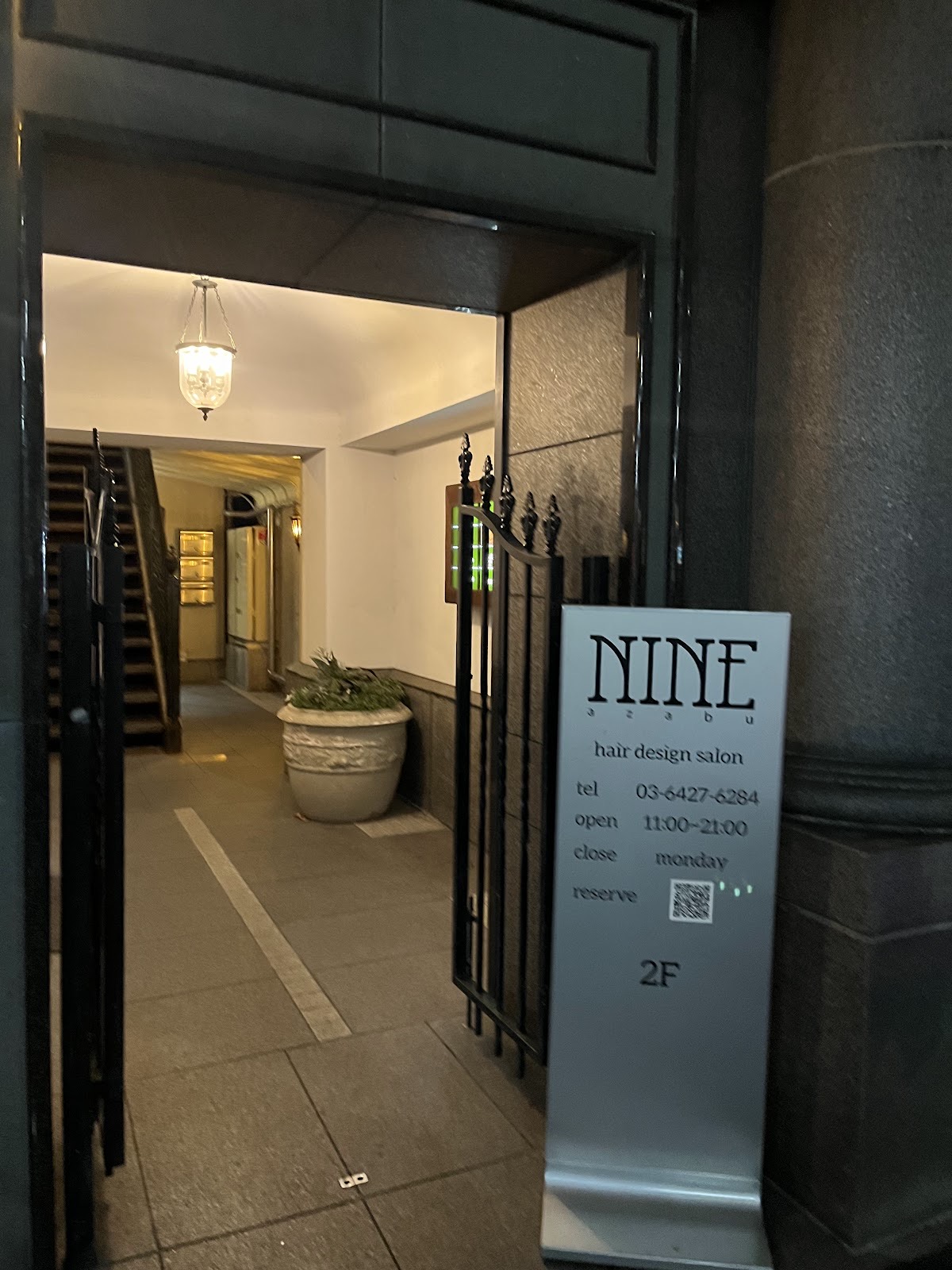 夜の入口。黒い鉄の門扉と磨かれた黒い石柱の間に、「NINE azabu / hair design salon / 2F」と書かれた淡い青灰色の立て看板。奥に真鍮のランタンと階段のある白いロビー。