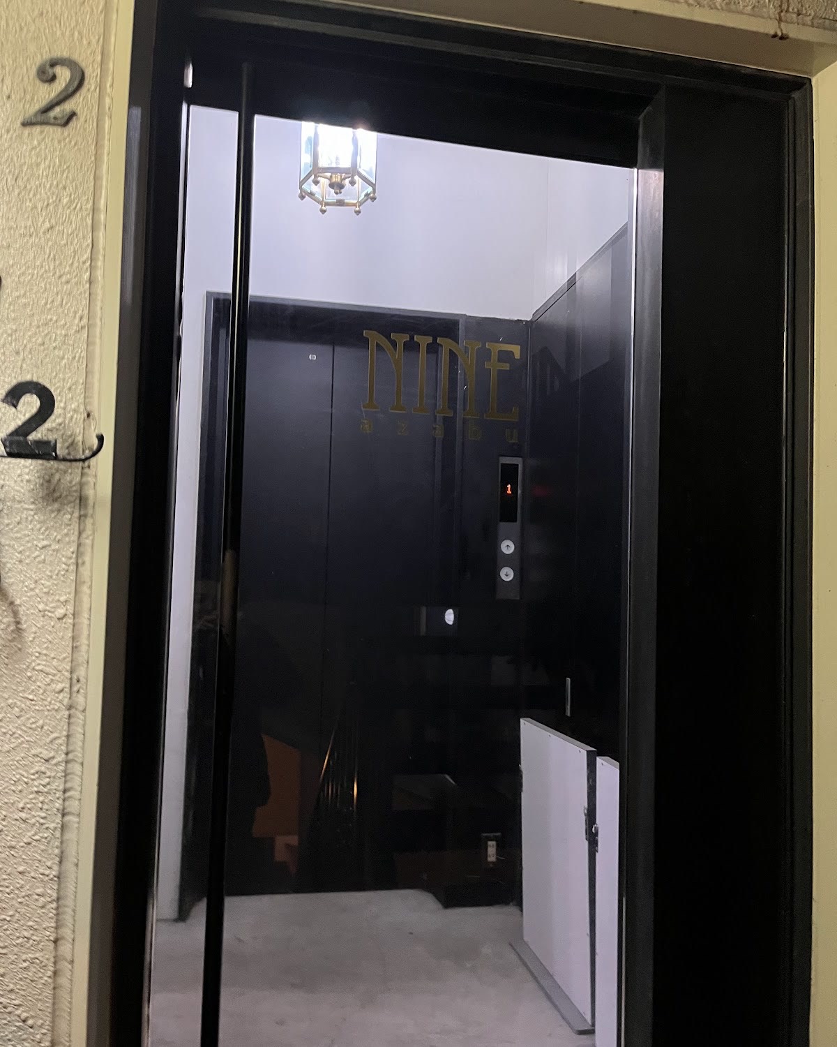 黒い枠のガラス扉越しに見える黒い壁。金色の「NINE azabu」のロゴ、六角形の真鍮ランタン、「1」と表示されたエレベーターのボタン。外壁には真鍮の「2」の数字。