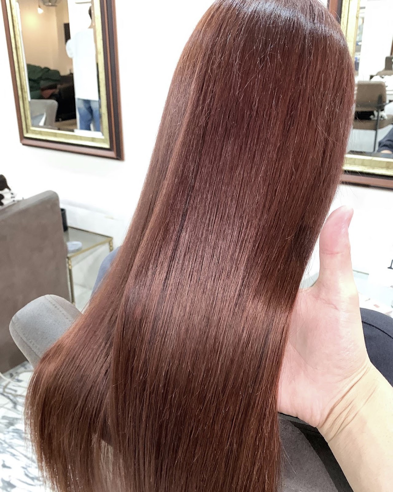 手で広げた深いピンクレッドの艶やかなストレートロング。白い壁に金縁の鏡、左にトープ色の椅子。