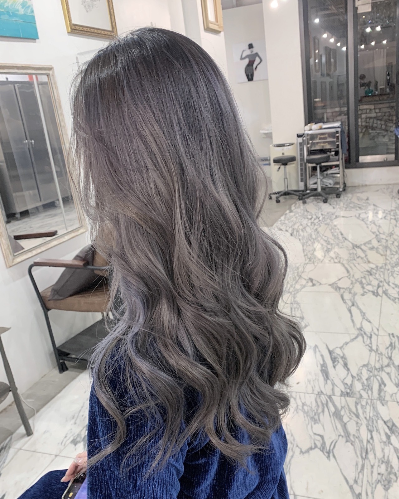 淡いシルバーグレーに染めた、ゆるく巻いたロングヘア。ロイヤルブルーのベルベットの服の後ろ姿、周りに白縁の鏡と白い大理石の床。