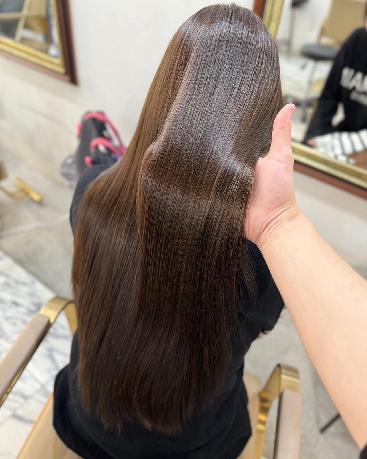 手で持ち上げた、光の筋が流れる艶やかなダークブラウンのロング。黒い服で真鍮枠の椅子に座る後ろ姿、右に金縁の鏡。