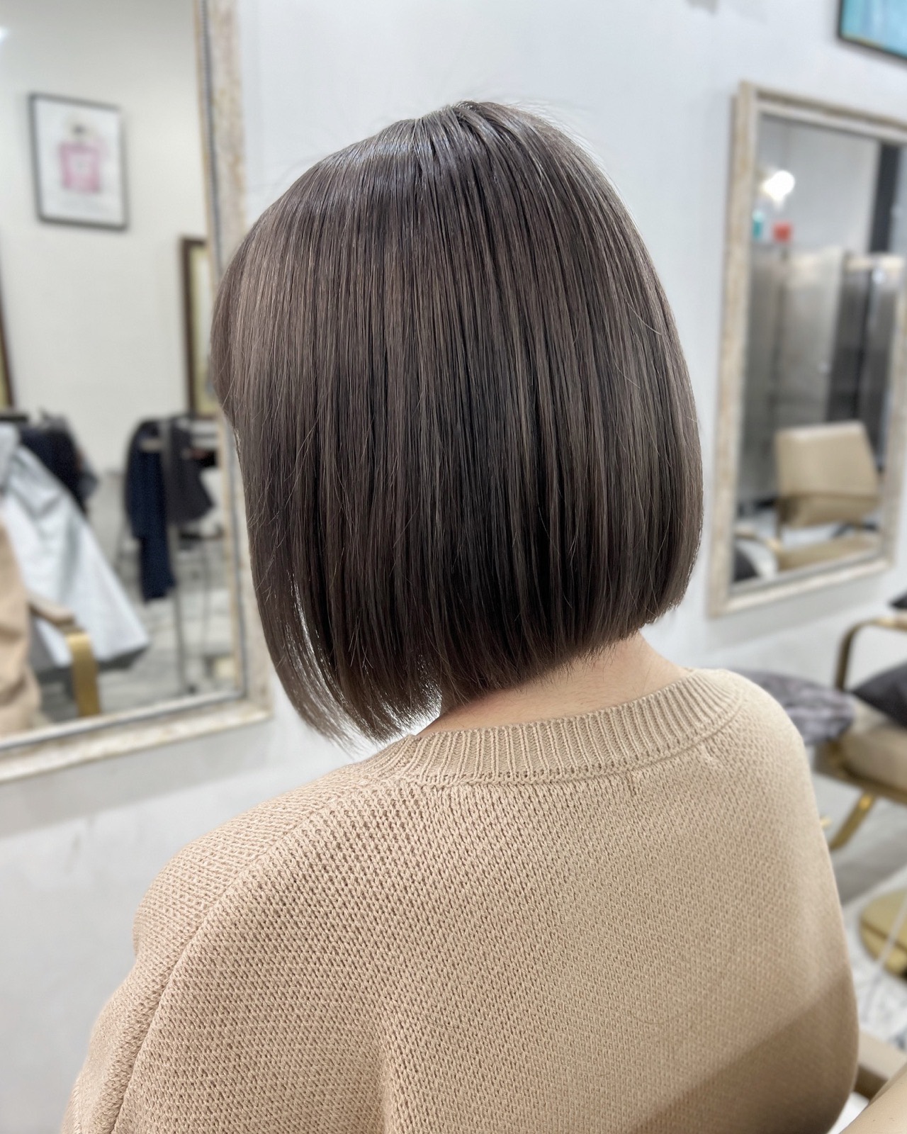 グレージュの丸みのあるボブ、ベージュのニットの後ろ姿。奥に白縁の鏡。