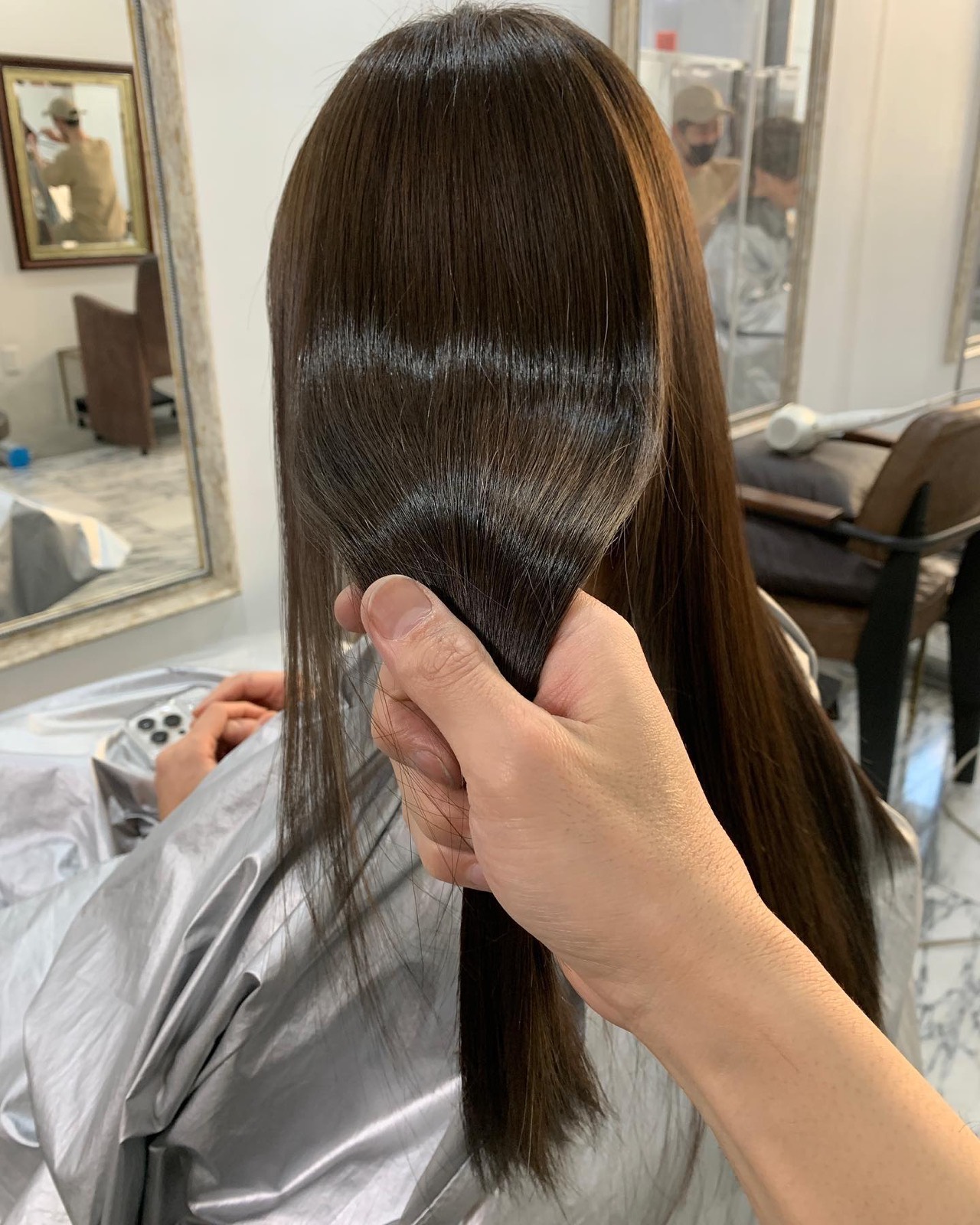 手で束ねた、幅広い艶の帯が出るダークブラウンのロング。シルバーのクロス、奥に鏡とタンカラーの椅子。